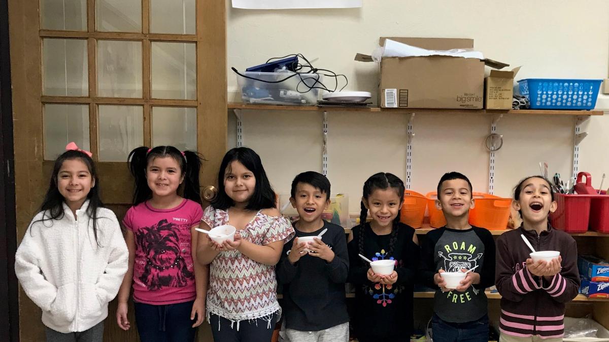 A diverse group of seven happy children holding bowls in a classroom, with a notice board and colorful storage shelves in the background.